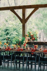 Wedding Planned by Terran Ward Events View of long-wooden reception table set up outside. Features red napkins black wired chairs, and tropical inspired florals. Wedding planned by luxury Asheville wedding planner Terran Ward