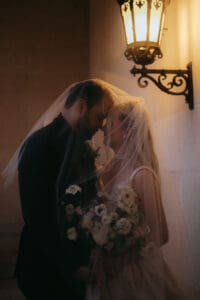 Biltmore Estate Asheville Wedding_Heather + Ben Close-up of bride and groom leaning their foreheads against each other under the bride's veil in the corner of a hallway. The bride carries a light colored, muted bouquet and they stand under an elegant lamp protruding from the wall. Wedding planned by luxury Asheville wedding planner Terran Ward