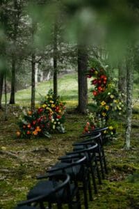 Wedding planned by Terran Ward Close-up view of outside wedding ceremony aisle and one side of guest seating. Guest seating are black wired chairs and the end of the aisle features half a floral arch with tropical inspired florals. Wedding planned by luxury Asheville wedding planner Terran Ward