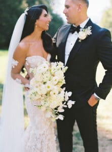 Biltmore Weddings – Marcos and Gaby Bride in lace gown with white bouquet smiling at groom in tuxedo outdoors at Biltmore Estate.