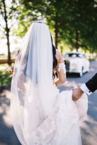 Biltmore Weddings – Marcos and Gaby Bride in long veil and lace gown walking toward vintage white car, groom holding her dress.