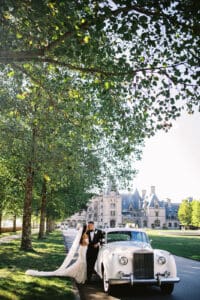 Biltmore Weddings – Marcos and Gaby Couple embracing beside a classic white car with the Biltmore Estate in the background.