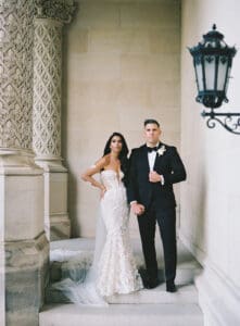 Biltmore Weddings – Marcos and Gaby Bride in lace gown and groom in tuxedo posing under ornate stone architecture.
