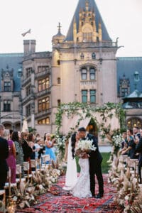 Biltmore Weddings – Marcos and Gaby Bride and groom share a romantic kiss at their outdoor wedding ceremony at the Biltmore Estate, surrounded by guests and floral aisle décor.