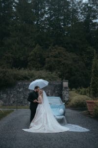 Biltmore Weddings – Heather + Ben Bride and groom kiss in front of a classic car, under an umbrella on the Biltmore Estate grounds.