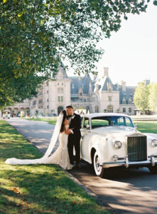Biltmore Estate Asheville Wedding_Marcos and Gaby Couple kissing beside a classic white car with the Biltmore Estate in the background. Wedding planned by Asheville wedding planner Terran Ward — bold editorial mountain wedding design.