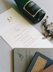 Biltmore Estate Asheville Wedding_Marcos and Gaby Wedding invitation with rings, champagne bottle, and books styled flat lay. Wedding planned by Asheville wedding planner Terran Ward — bold editorial mountain wedding design.