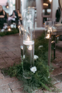 Daniela and Angelo Wedding Grand Bohemian Asheville Close up of aisle decoration for an outside ceremony under a tent featuring greenery and candles in long, tall glass containers. Wedding Planned by Terran Ward at The Grand Bohemian Asheville