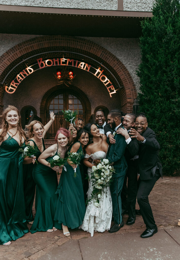 Couple posing happily with bridesmaids, in dark green, and groomsmen in front of the bohemian hotel. Wedding planned by Terran Ward