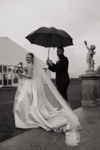 Biltmore Estate Asheville Wedding_Heather + Ben Black and white photo of groom holding umbrella for bride holding a bouquet outside the Biltmore Estate. A tent is in the background. Wedding rain plan planned by Asheville wedding planner Terran Ward.