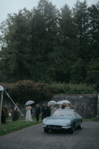 Biltmore Estate Asheville Wedding_Heather + Ben Couple and wedding guests gathered under umbrellas by a classic blue car outside the Biltmore Estate. The edge of a tent can be seen in the middle ground. Wedding rain plan planned by Asheville wedding planner Terran Ward.