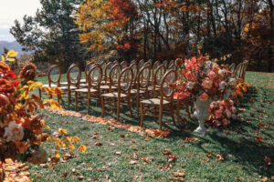 Mountain Wedding – Kaitlyn and Eric Side view of outdoor wedding ceremony aisle featuring light wooden chairs, bold fall florals, and one large bouquet in a stone vase. Wedding planned by Asheville wedding planner Terran Ward — bold editorial design setup.