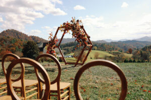 Mountain Wedding – Kaitlyn and Eric Wedding ceremony set up; light wooden chairs in the foreground, a hexagonal, wooden wedding arch with bold florals in the mid ground and mountains in the background. Wedding planned by Asheville wedding planner Terran Ward — bold editorial design setup.