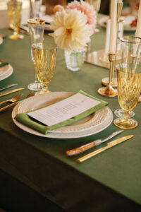 Mountain Wedding – Kaitlyn and Eric A close-up of an elegant table layout with gold-tinted glasses, a green table cloth, white plates, a green napkin, and gold-tinted silverware. Wedding planned by Asheville wedding planner Terran Ward — bold editorial design setup.