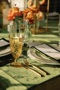 Mountain Wedding – Kaitlyn and Eric A close-up of an elegant table layout with gold-tinted glasses, a green table cloth, white plates, a green napkin, and gold-tinted silverware. Wedding planned by Asheville wedding planner Terran Ward — bold editorial design setup.