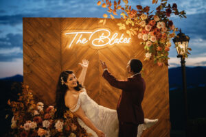 Mountain Wedding – Kaitlyn and Eric Bride and groom gesturing to the background of a photo op set-up. The background is wooden with a light-up board saying "The Blakes." Wedding planned by Asheville wedding planner Terran Ward — bold editorial design setup.