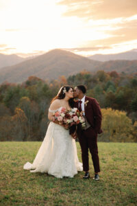 bride and groom kiss in front of mountain sunset, groom in a maroon suite and bride in a floral white gown. Asheville wedding planner Terran Ward created sunset-driven timeline for mountain wedding