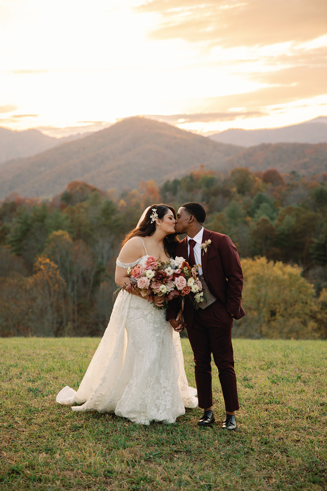 bride and groom kiss in front of mountain sunset, groom in a maroon suite and bride in a floral white gown. Asheville wedding planner Terran Ward created sunset-driven timeline for mountain wedding