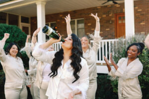 Asheville Mountain Wedding_Kaitlyn and Eric Bride celebrating with her bridesmaids, in silk pajamas, in the morning of the wedding by drinking form a champagne bottle outside. Wedding planned by Asheville wedding planner Terran Ward — bold editorial mountain wedding design.