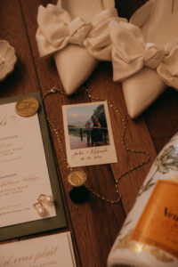 Chestnut Ridge Asheville Wedding_Mikaela + Jacob Flat lay of wedding invitation, white heels with bows, a gold-chain necklace, and a polaroid of the couple. Wedding planned by Asheville wedding planner Terran Ward — bold editorial mountain wedding design.