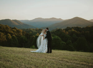 Couple kiss in front of sunset mountain background, bride in a floral lace white gown. Groom hugs the bride and the bride has her arms out wide. Asheville wedding planner Terran Ward created sunset-driven timeline for mountain wedding
