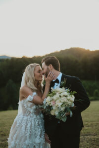 Bride and groom looking at each other, their noses touching and the bride's hand is on the groom's cheek in front of a sunset mountain background. The groom holds the bride's bouquet. Asheville wedding planner Terran Ward created sunset-driven timeline for mountain wedding