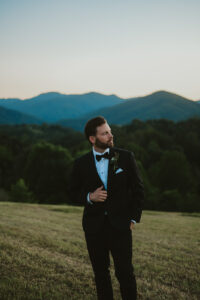 Groom glancing to the side in front of sunset mountain views in a black tuxedo. Asheville wedding planner Terran Ward created sunset-driven timeline for mountain wedding
