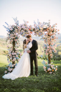 The Ridge Wedding Asheville Planned by Terran Ward Couple kissing under pastel floral wedding arch at The Ridge. Wedding planned by Asheville wedding planner Terran Ward — bold editorial mountain wedding design.