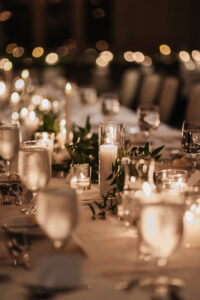 Daniela and Angelo Wedding Grand Bohemian Asheville Close up of a table set-up featuring candles in glass containers, green leaves, and clear glasses on top of a white table cloth. Wedding Planned by Terran Ward at The Grand Bohemian Asheville