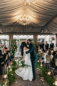 Daniela and Angelo Wedding Grand Bohemian Asheville Bride and groom kissing in the aisle under a tent outside with chandeliers. Wedding guests clap and cheer. Wedding Planned by Terran Ward at The Grand Bohemian Asheville