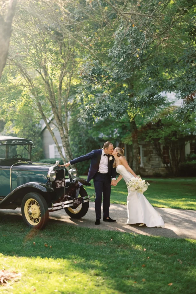 Bride leaning over to kiss groom next to old green car at the farm at Old Edwards. Wedding planned by Terran Ward