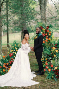Wedding Planned by Asheville Planner Terran Ward Bride and groom holding hands and smiling towards the camera in front of their floral arch with tropical themed flowers. Wedding planned by Asheville wedding planner Terran Ward — bold editorial design setup.
