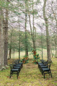 Darrell – April Styled Shoot Wide view of outdoor wedding ceremony set-up in the woods featuring a tropical-themed floral wedding arch, and black framed chairs.