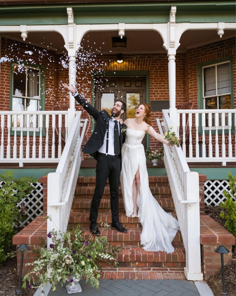 Bride and groom smiling on the steps outside of Zelda Dearest, as the groom throws flower petals in. the air.
