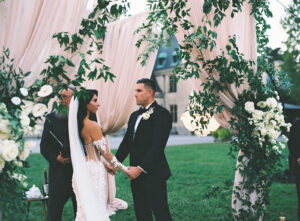 Couple standing in front of officiant under a white flower and cream-pink fabric wedding arch. Destination wedding in Asheville — Terran Ward planning mountain ceremony