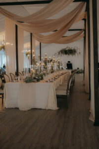 Long reception table featuring white linens, classic chairs, candles, and light florals under neutral draping. Asheville wedding planner Terran Ward creating luxury weddings with a budget.