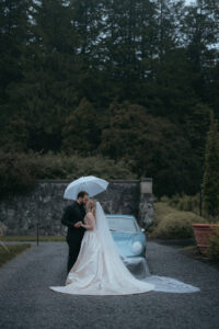 Couple kissing in front of a classic light blue car under an umbrella on rainy day. Destination wedding in Asheville — Terran Ward planning mountain ceremony