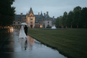 Back view of bride and groom walking to the Biltmore Estate in the rain, under an umbrella. Destination wedding in Asheville — Terran Ward planning mountain ceremony
