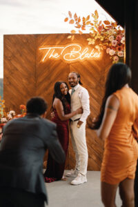 Wedding guests happily posing in front of photo backdrop featuring a wooden background, fall florals, and a lit up sign saying "The Blakes." Asheville wedding planner Terran Ward creating luxury weddings with a budget.