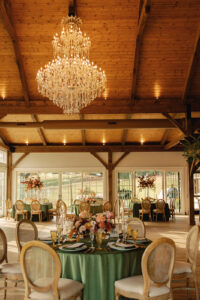 Portrait view of reception room featuring a chandelier, and circular tables with green tableclothes and fall florals. Asheville wedding planner Terran Ward creating luxury weddings with a budget.