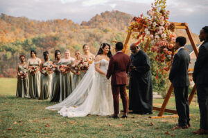 bride and groom holding hands and smiling in front of officiant. Bridesmaids in green stand behind the bride and we can see a few of the groomsmen behind the groom. Destination wedding in Asheville — Terran Ward planning mountain ceremony.