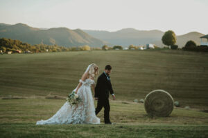 Landscape view of bride and groom walking, holding hands, with rolling mountains in the background and haybales. Asheville micro wedding — modern editorial setup by Terran Ward
