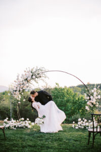 Groom dipping and kissing bride under a floral wedding arch at Serenity Ridge. Asheville micro wedding — modern editorial setup by Terran Ward